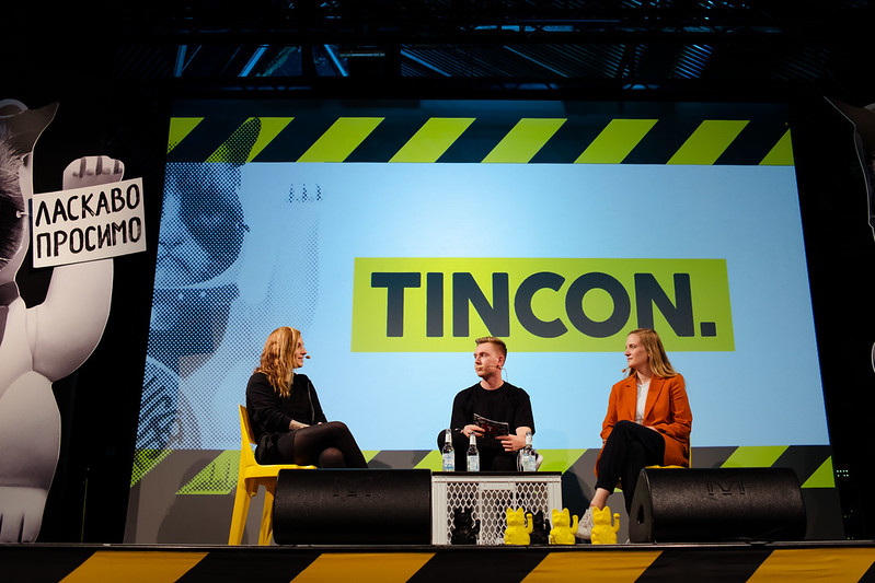 Zu sehen ist eine Bühne. Im Hintergrund, das Logo der TINCON. Im Vordergrund sitzen drei Personen, sie sind sich im Gespräch zugewandt sind. Bei den Sprecher*innen handelt es sich um Isabell Beer, Tomatolix und Carmen Wegge.