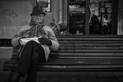 Auf dem Bild ist ein Mann, der auf einer Bank sitzt, grimmig schaut und Zeitung in der hand hält.