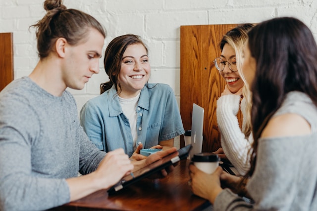 Vier junge lachende Personen arbeiten am Laptop mit Kaffee.