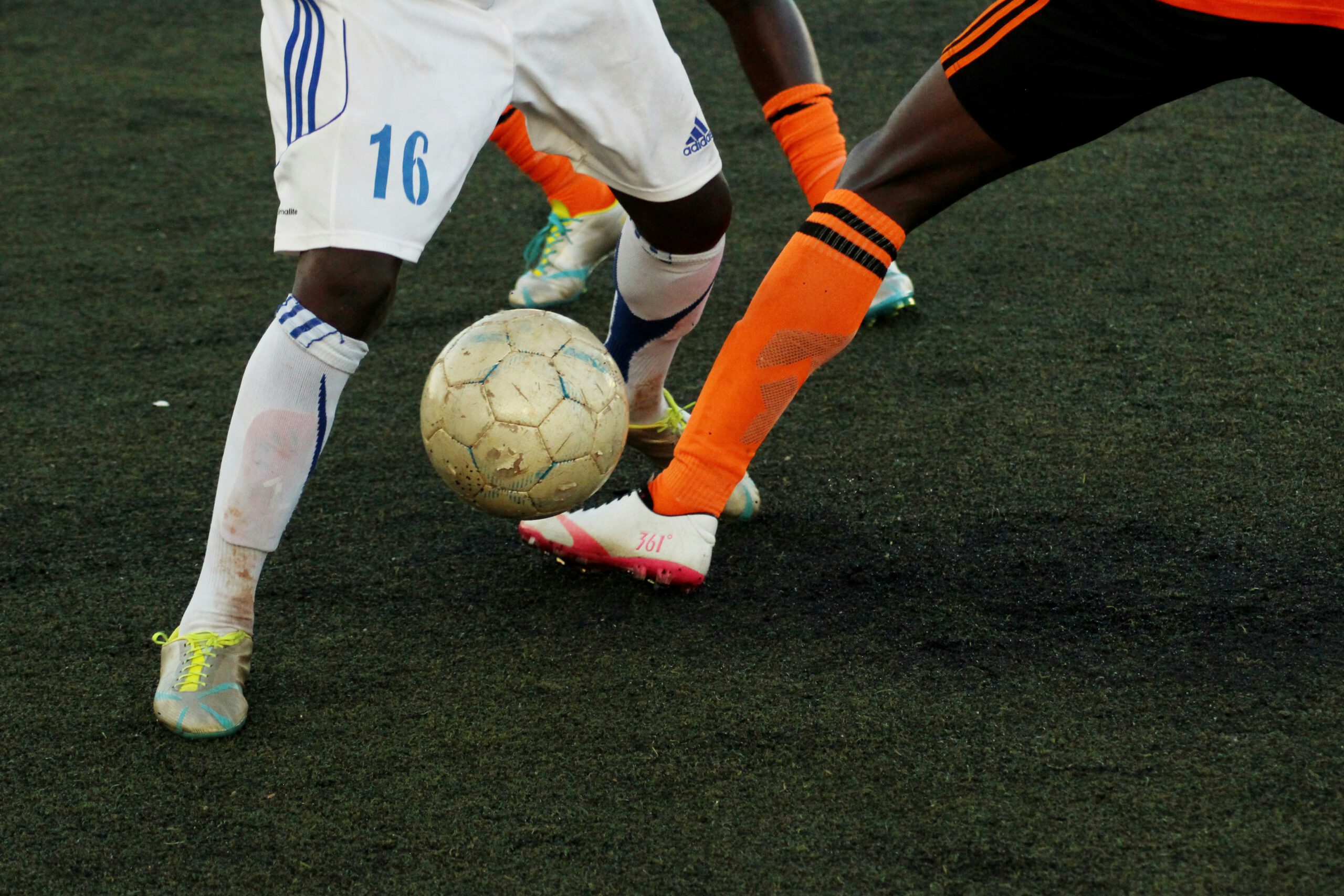 Die Beine zweier Fußballspieler im Kampf um den Ball. Einer trägt orangefarbene Strümpfe, sein Gegener trägt weiß.